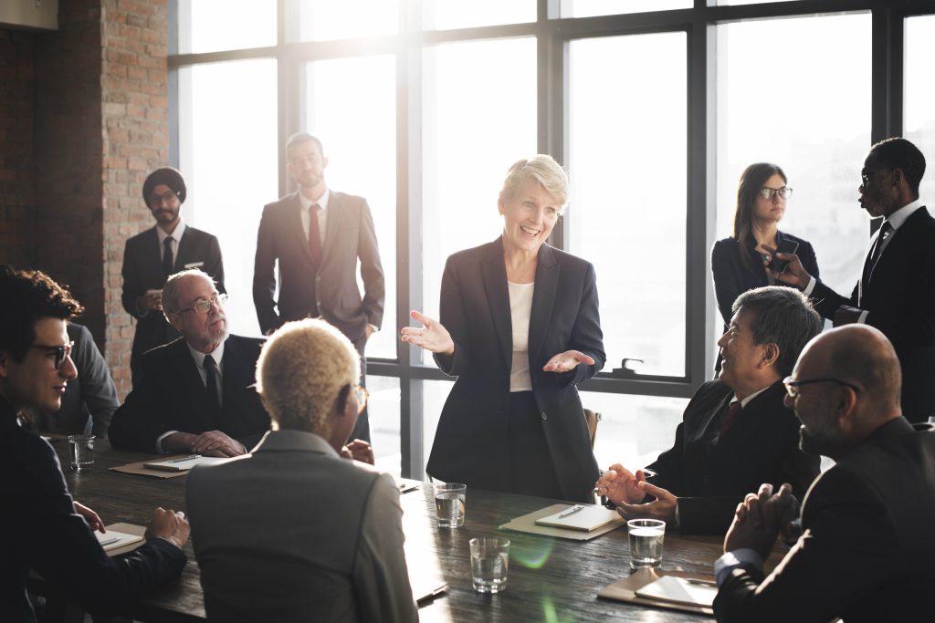 Woman presenting to business people around a conference table by tall windows