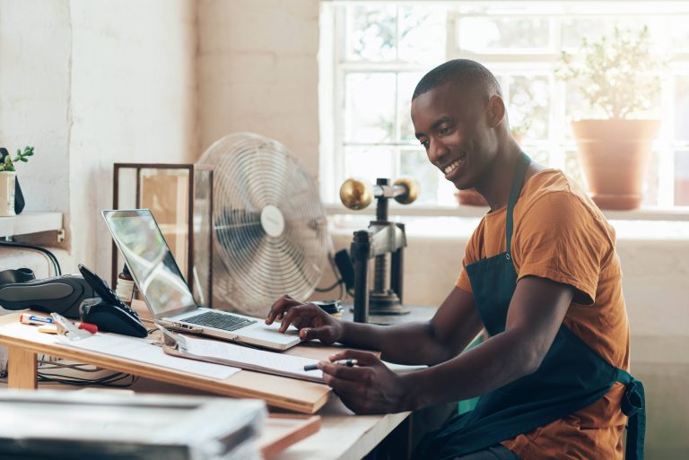 Smiling small business owner in an apron typing on a laptop in a workshop