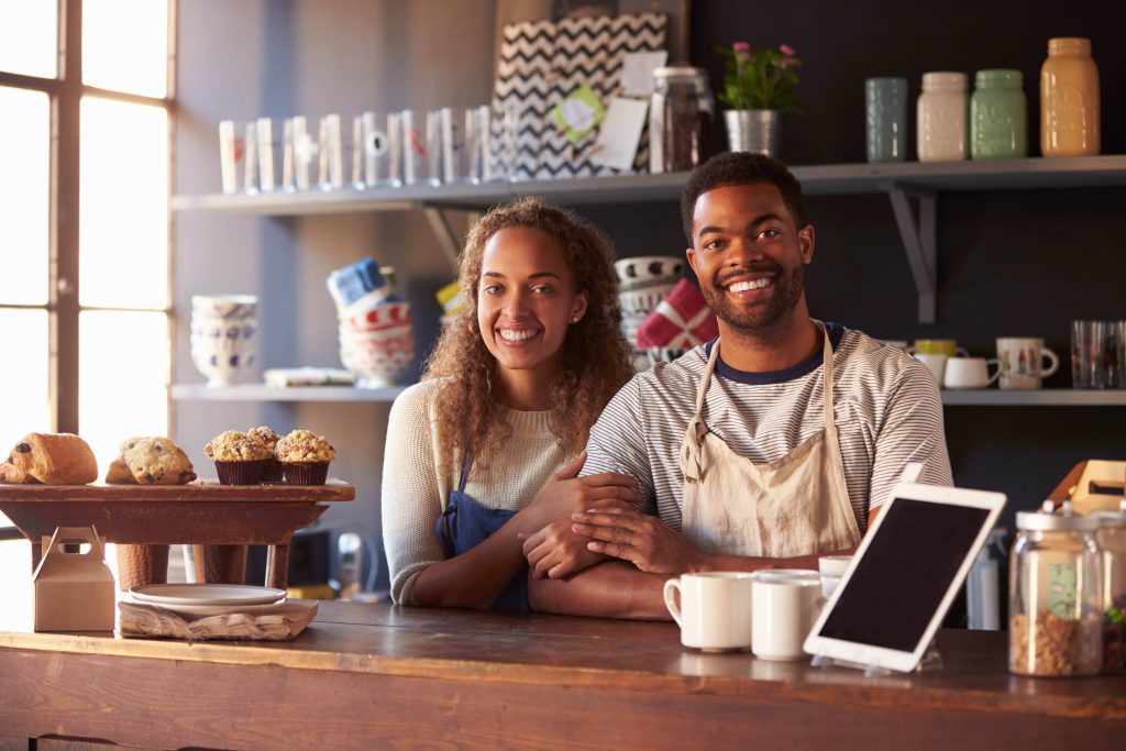 Smiling cafe owners leaning on the counter beside pastries and a tablet