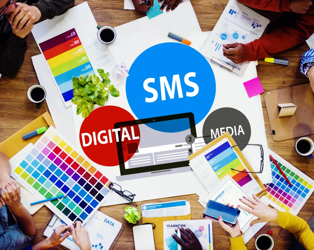 Team around a table with color swatches and bubbles reading SMS, Digital and Media