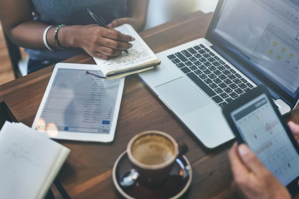 Desk with a laptop, tablet, phone and coffee while a person writes notes