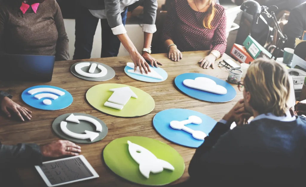 Team around a wooden table holding colorful circular icons for wifi, cloud and launch