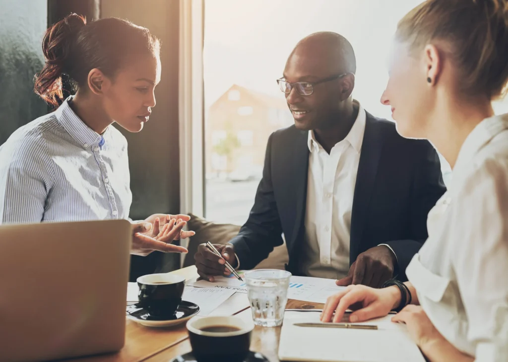 Three colleagues talking at a table with a laptop and coffee in a bright office