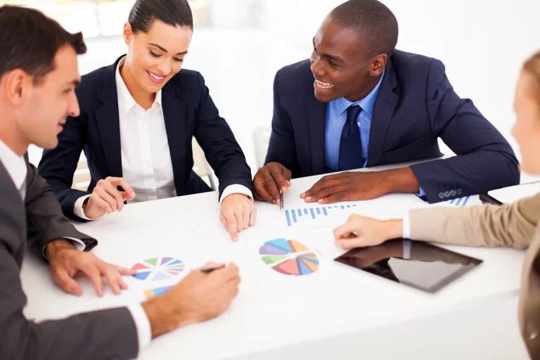 Four business people at a white table reviewing pie charts and a tablet