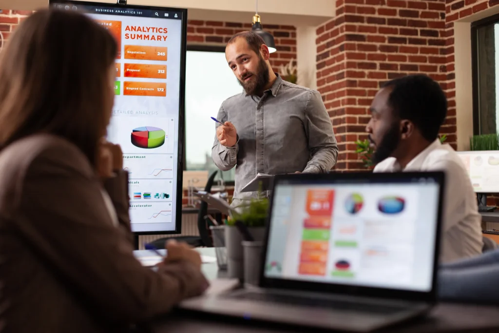 Man presenting charts on a screen to colleagues in a brick-walled office
