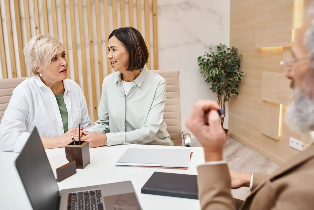 Three women talking at a table with a laptop in a wood-paneled office