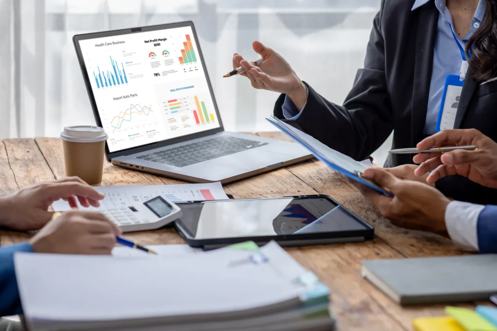 Colleagues at a wooden table pointing at charts on a laptop beside a tablet and coffee