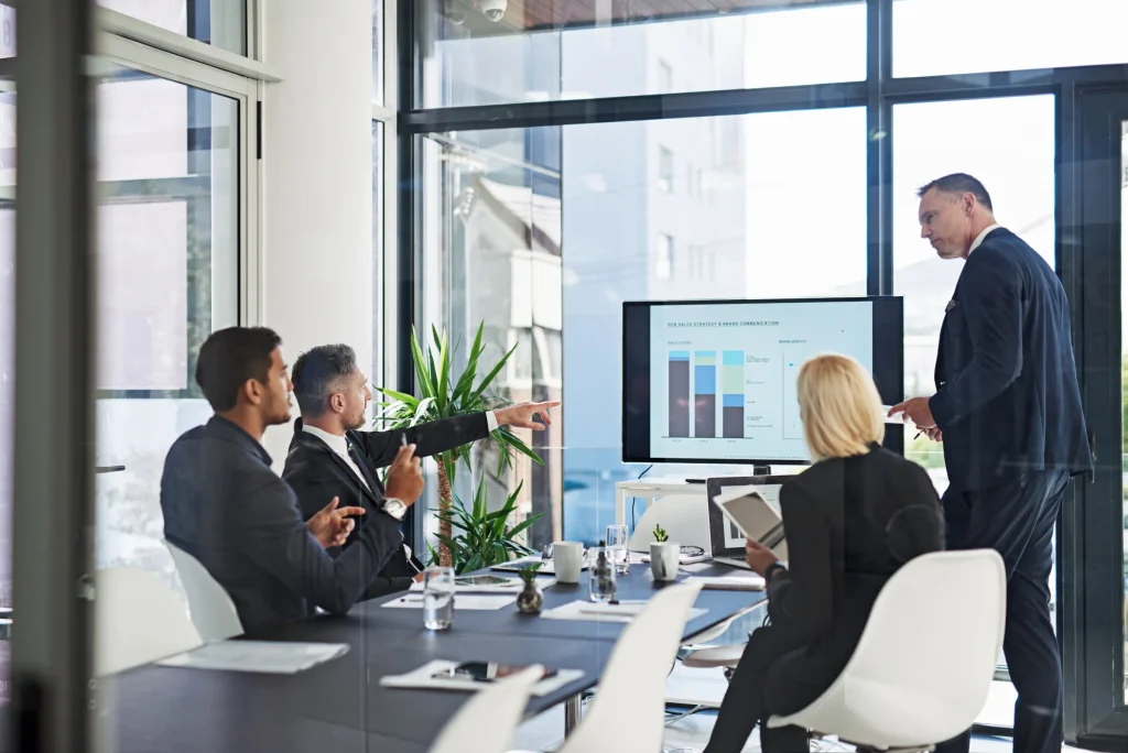 Team in a glass-walled meeting room while a man presents a chart on a screen