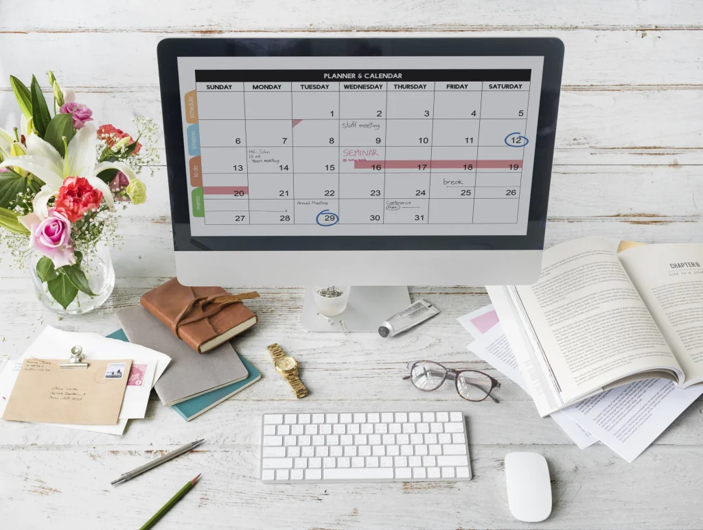 Desktop computer showing a calendar planner on a white desk with flowers and glasses