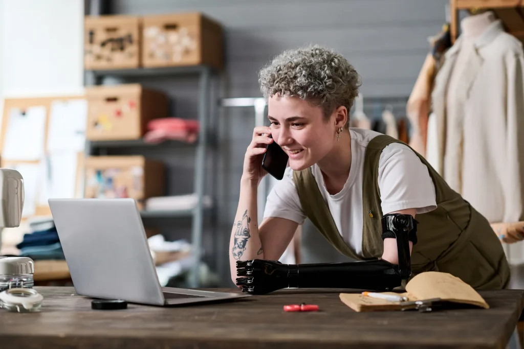 Smiling woman talking on the phone at a laptop in a clothing workshop